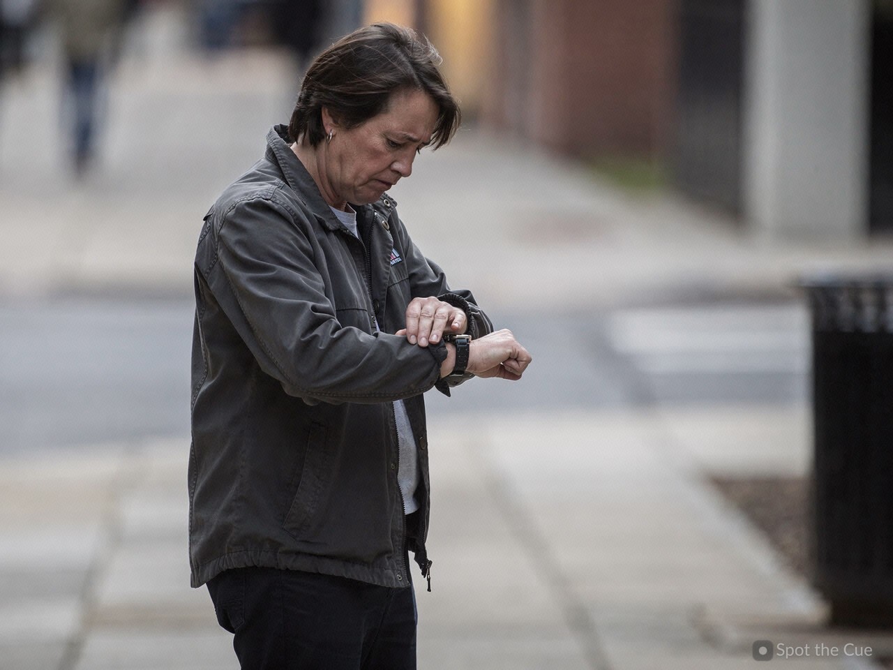 A person in a jacket glancing down at their wristwatch with a slightly tense, expectant expression.