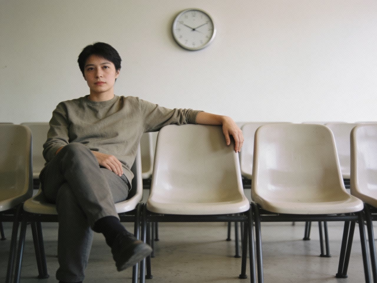 A person sitting in a row of hard molded plastic chairs in a bare waiting room, mid-shift in their seat with legs re-crossing, a clock on the wall behind.